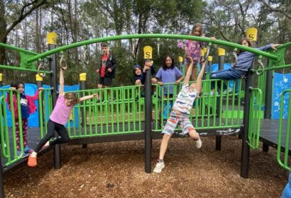 A group of children plays on a green and black playground structure surrounded by trees. Some children are climbing, while others are hanging or standing on the play equipment.