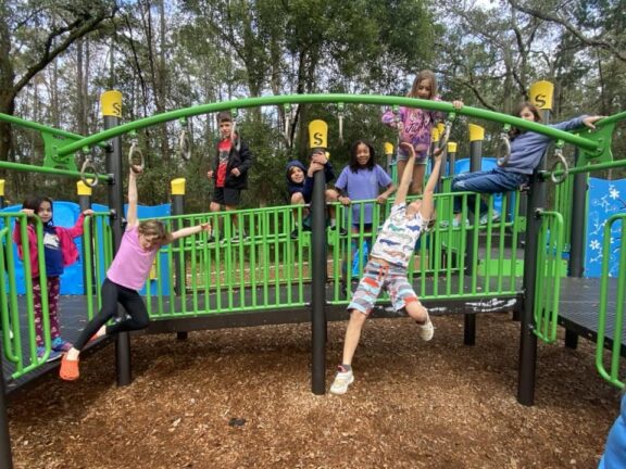 A group of children plays on a green and black playground structure surrounded by trees. Some children are climbing, while others are hanging or standing on the play equipment.