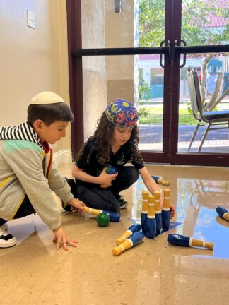 Two children are playing with a wooden bowling set on the floor indoors. The child on the left is wearing a kippah and a jacket, and the child on the right is wearing a colorful head covering.