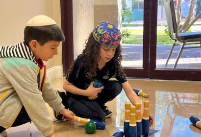 Two children are playing with a wooden bowling set on the floor indoors. The child on the left is wearing a kippah and a jacket, and the child on the right is wearing a colorful head covering.