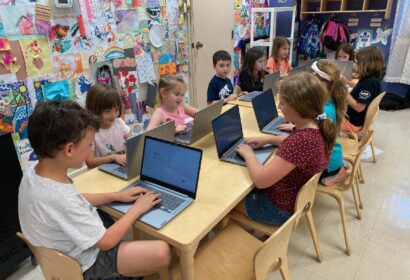 A group of young children seated at a classroom table, each using a laptop. The background shows colorful art displayed on the walls and backpacks hung on hooks.