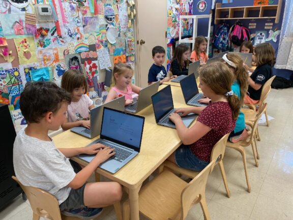 A group of young children seated at a classroom table, each using a laptop. The background shows colorful art displayed on the walls and backpacks hung on hooks.