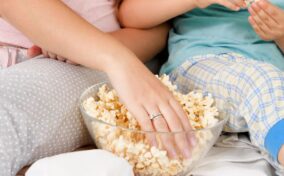 Two people sitting close together, reaching into a bowl of popcorn. One person wears pajamas with gray polka dots, and the other person wears plaid pajamas in light blue and yellow.