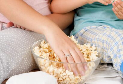 Two people sitting close together, reaching into a bowl of popcorn. One person wears pajamas with gray polka dots, and the other person wears plaid pajamas in light blue and yellow.