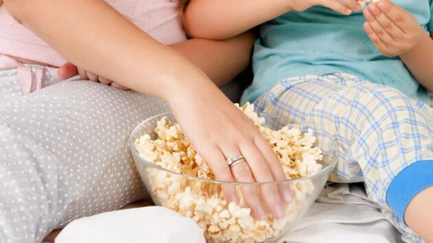 Two people sitting close together, reaching into a bowl of popcorn. One person wears pajamas with gray polka dots, and the other person wears plaid pajamas in light blue and yellow.