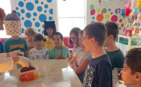 A group of children stand around a table watching a person in white gloves handle items. The room is brightly decorated with polka dots, colorful objects, and hanging decorations.