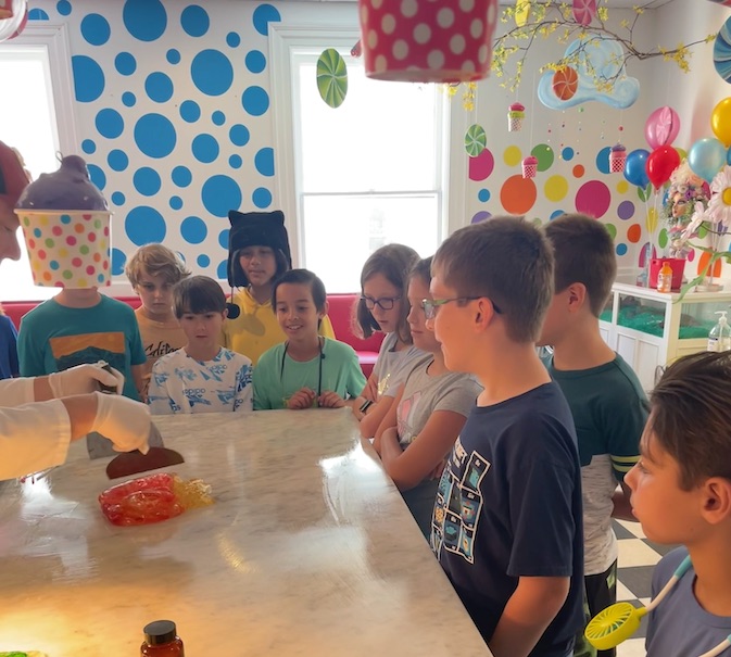 A group of children stand around a table watching a person in white gloves handle items. The room is brightly decorated with polka dots, colorful objects, and hanging decorations.