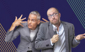 Two men in gray blazers stand in front of a blue background with geometric patterns. One man gestures animatedly with his hands, while the other holds a microphone and gestures with a shrug.