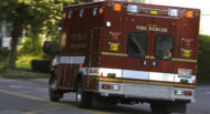 A fire rescue ambulance with emergency lights on is driving through a suburban street. The vehicle is red and white, marked with "Fire Rescue" on the back. Trees and buildings are in the background.