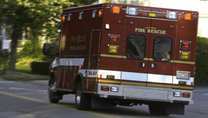 A fire rescue ambulance with emergency lights on is driving through a suburban street. The vehicle is red and white, marked with "Fire Rescue" on the back. Trees and buildings are in the background.