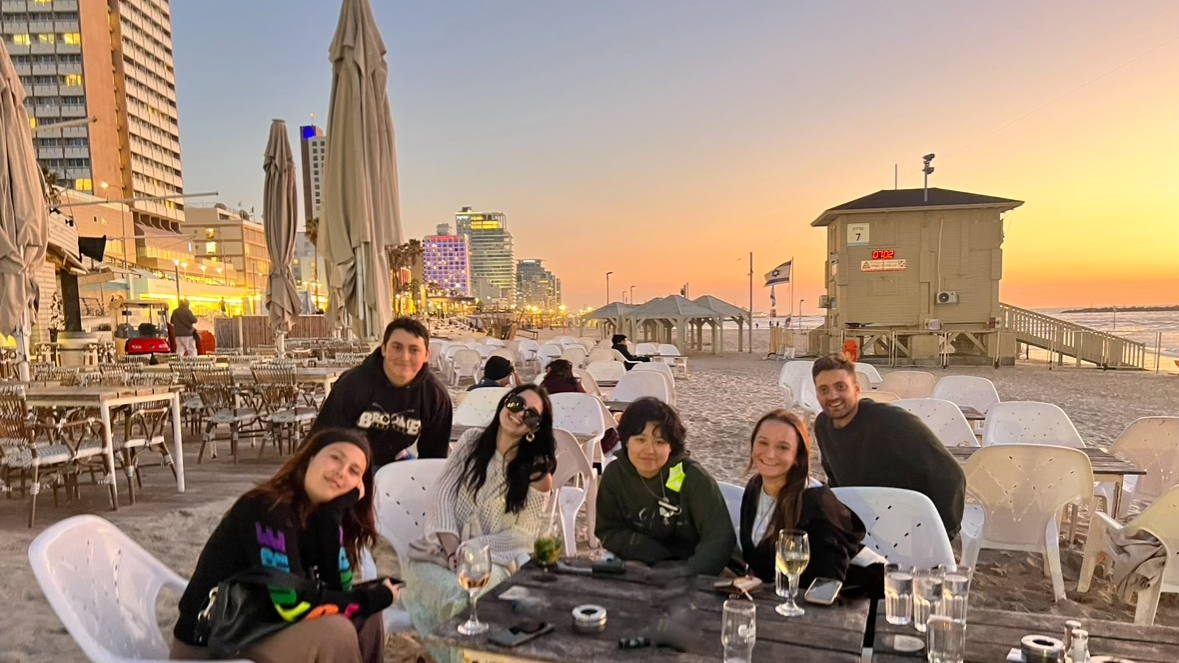 A group of six people sitting around a table on a sandy beach, with buildings and a sunset in the background.