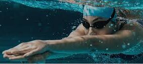 A swimmer in a pool underwater, wearing a blue swim cap and goggles, extends their arms forward.