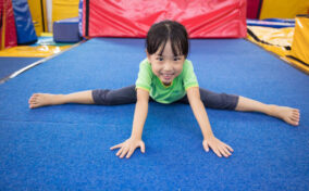 Child in a green shirt and gray pants stretching in a gymnasium on a blue mat, smiling at the camera.