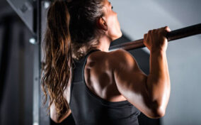 A woman with long hair in a ponytail performs a pull-up on a metal bar. She is wearing a black sports bra.