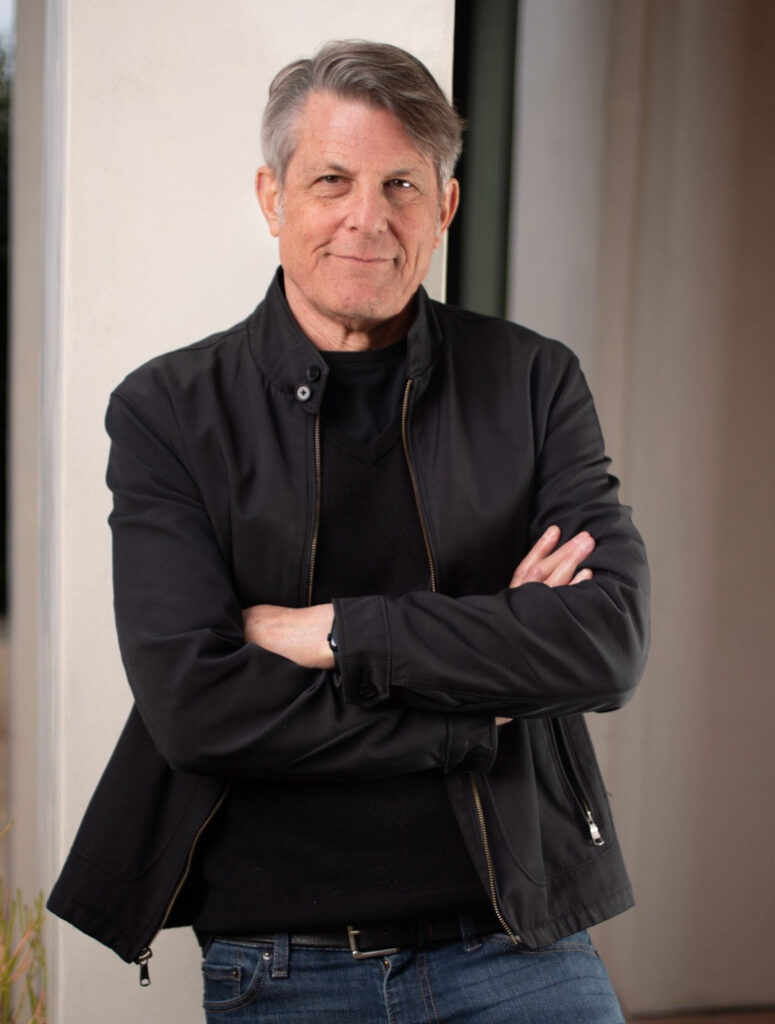 A person with short gray hair and a black jacket stands with arms crossed in front of a white wall.