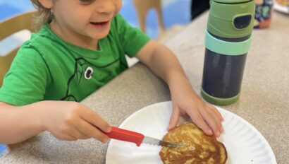 Child in a green shirt uses a red knife to spread on a pancake at a table. A green water bottle is beside the plate. Another child appears in the background.