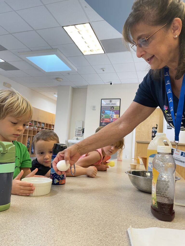 A woman assists a group of children with an egg during a kitchen activity at a table.