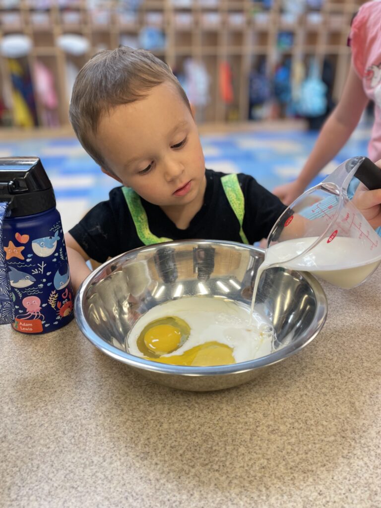 A child pours liquid into a metal bowl containing eggs, with a water bottle nearby on the table.