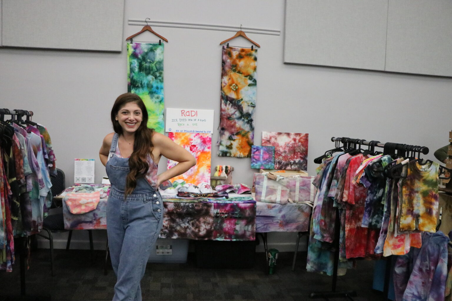 A woman stands in front of a display of colorful tie-dye clothing and fabric art at a booth.