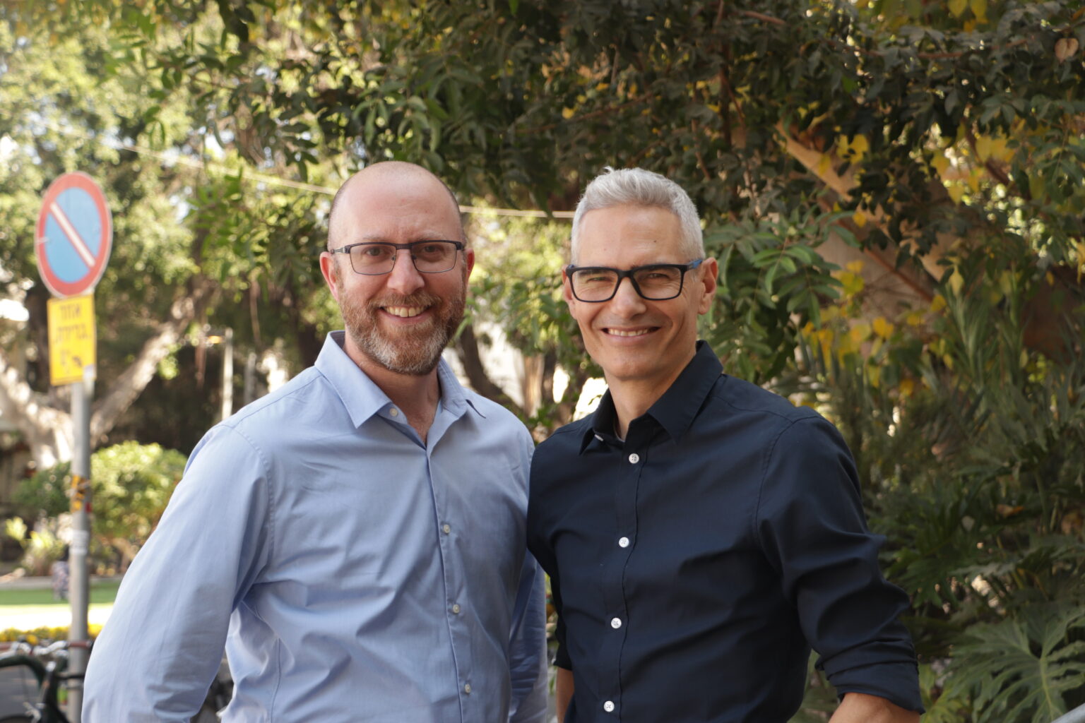 Two people standing outdoors in front of trees, smiling at the camera.