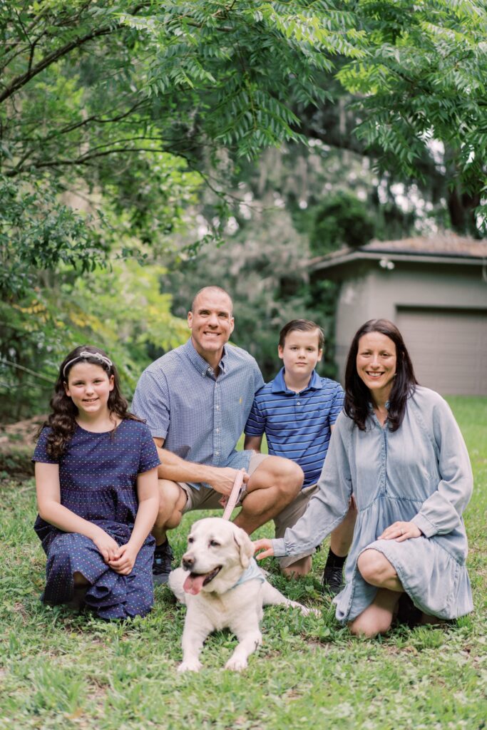 A family of four kneels on the grass with a white dog. Two adults and two children are dressed in blue, and trees and a shed are in the background.