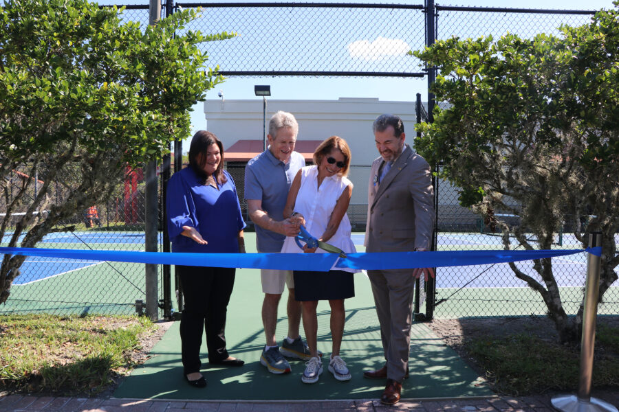 Four people stand at a tennis court ribbon-cutting ceremony, with one person holding large scissors to cut the blue ribbon.