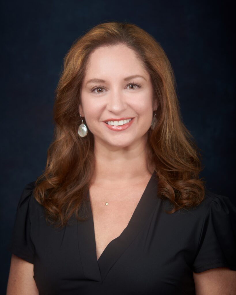 A woman with long brown hair, wearing a black blouse and earrings, smiles in front of a dark background.