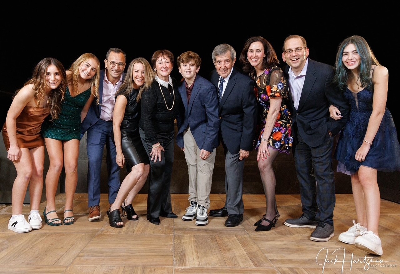 A group of ten people posing together in formal and semi-formal attire on a wooden floor, in front of a black background.