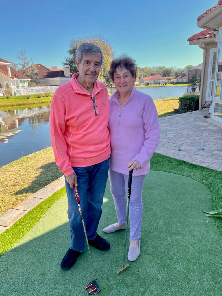 Two people stand on a putting green, holding golf clubs. They are outdoors near a pond with a clear sky and houses in the background.