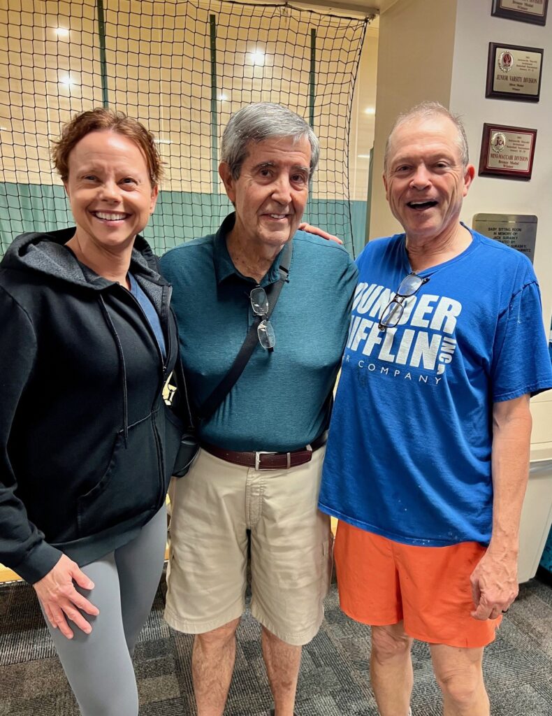 Three people standing indoors, smiling at the camera. The person in the middle has a sling on their arm. The other two are wearing casual clothing. Plaques are visible on the wall behind them.