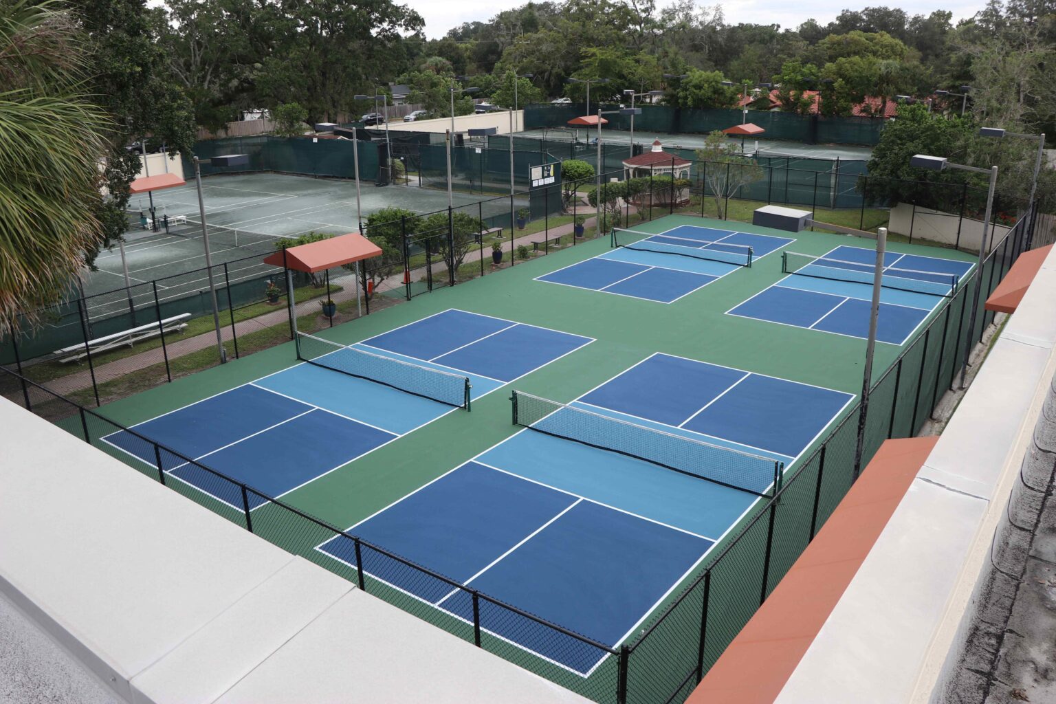 An aerial view of a set of blue and green outdoor tennis and pickleball courts, surrounded by fencing, with trees and additional tennis courts in the background.