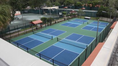 An aerial view of a set of blue and green outdoor tennis and pickleball courts, surrounded by fencing, with trees and additional tennis courts in the background.