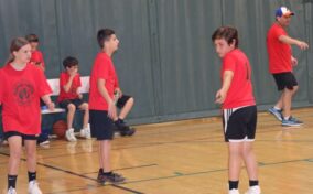 A youth basketball team in red jerseys on the court during a game, with a coach in the background.