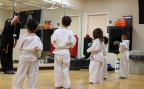 Children in white martial arts uniforms stand in a line facing an instructor in a gym with mirrored walls.