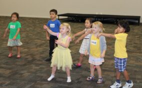 Six children stand on a carpeted floor, facing forward, as if responding to instructions.