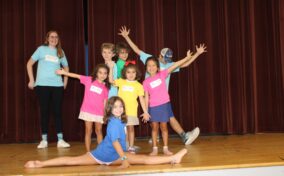 Seven children and one adult pose happily on a stage. A girl in front does the splits while others stand and strike poses. They're wearing colorful shirts with name tags.