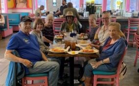 A group of seven adults seated around a table in a colorful diner, with food and drinks in front of them.