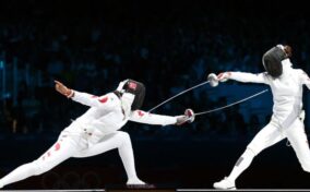 Two fencers in white protective gear compete in a match, each holding a foil.