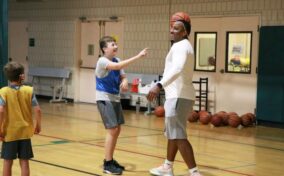 A basketball coach balances a ball on his head while kids in sports attire stand around, smiling in a gym.