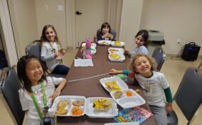 Five children sitting around a table having a meal, with various food items like sandwiches and fruit on trays. They are smiling and wearing name tags.