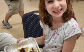 A smiling girl holds a tray with three decorated pastries, topped with sprinkles, in a room with other people in the background.