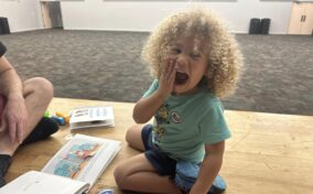 Child with curly hair sits on a wooden floor, holding a hand to their mouth. They are surrounded by books and toys.