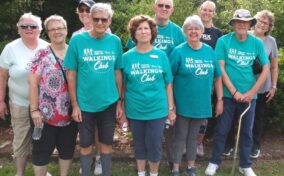 A group of older adults in "Walking Club" shirts stand together outdoors on grass, surrounded by trees.