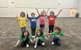 Eight children pose in colorful T-shirts with arms raised, standing and kneeling on a carpeted floor in a large room.