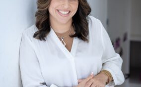 Woman in a white blouse leans against a wall, smiling. She has shoulder-length brown hair and is wearing a necklace and bracelet.