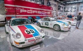 A display of vintage Porsche race cars, including a white car with "59" on the hood, in a museum setting. A large red truck with "Porsche" is in the background.