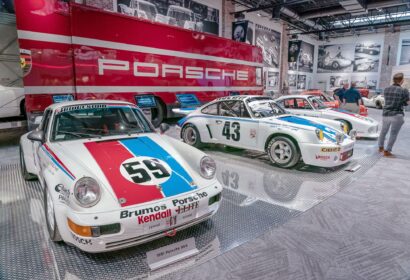 A display of vintage Porsche race cars, including a white car with "59" on the hood, in a museum setting. A large red truck with "Porsche" is in the background.