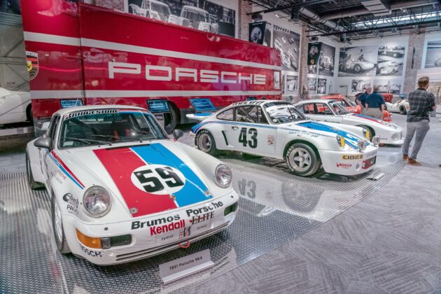 A display of vintage Porsche race cars, including a white car with "59" on the hood, in a museum setting. A large red truck with "Porsche" is in the background.