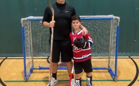 Two people pose in front of a small hockey goal indoors. The adult holds a hockey stick, while the child wears a red jersey and holds a ball.