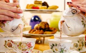 Two people pouring tea from floral teapots into matching cups, with a tiered tray of pastries in the background.